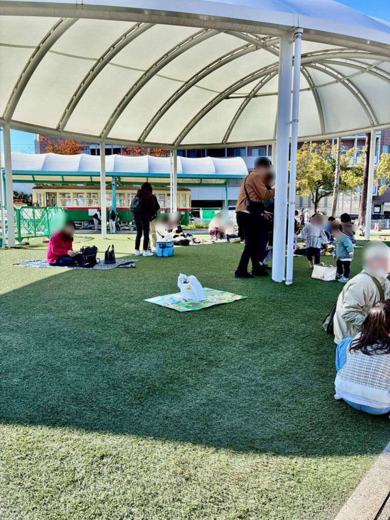 人工芝が敷かれた屋根付きの休憩スペースでレジャーシートを敷いてお昼ご飯を食べる家族連れ